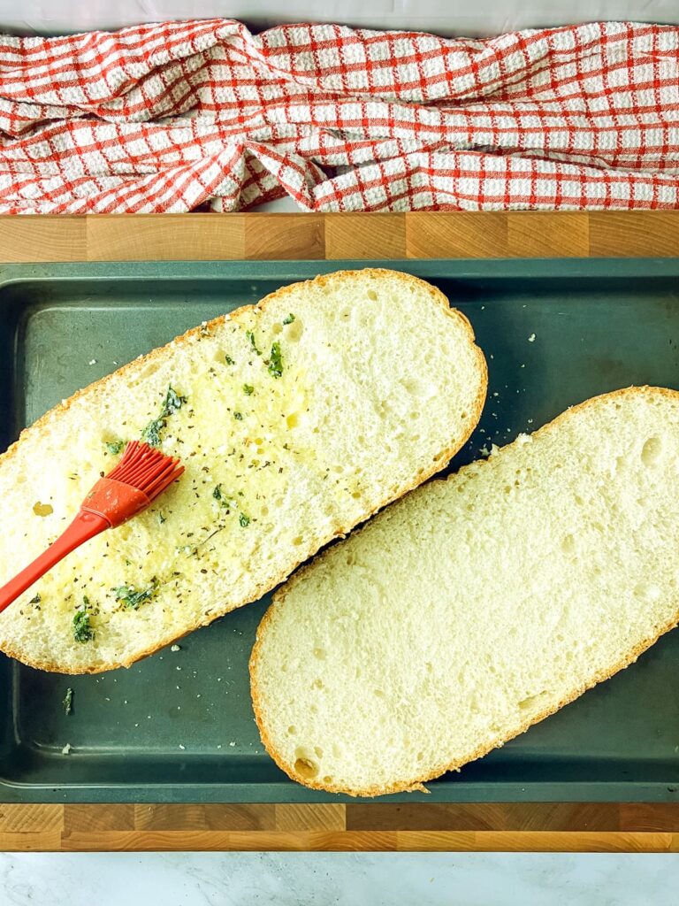A loaf of French bread cut in half on a baking tray being basted with garlic butter.