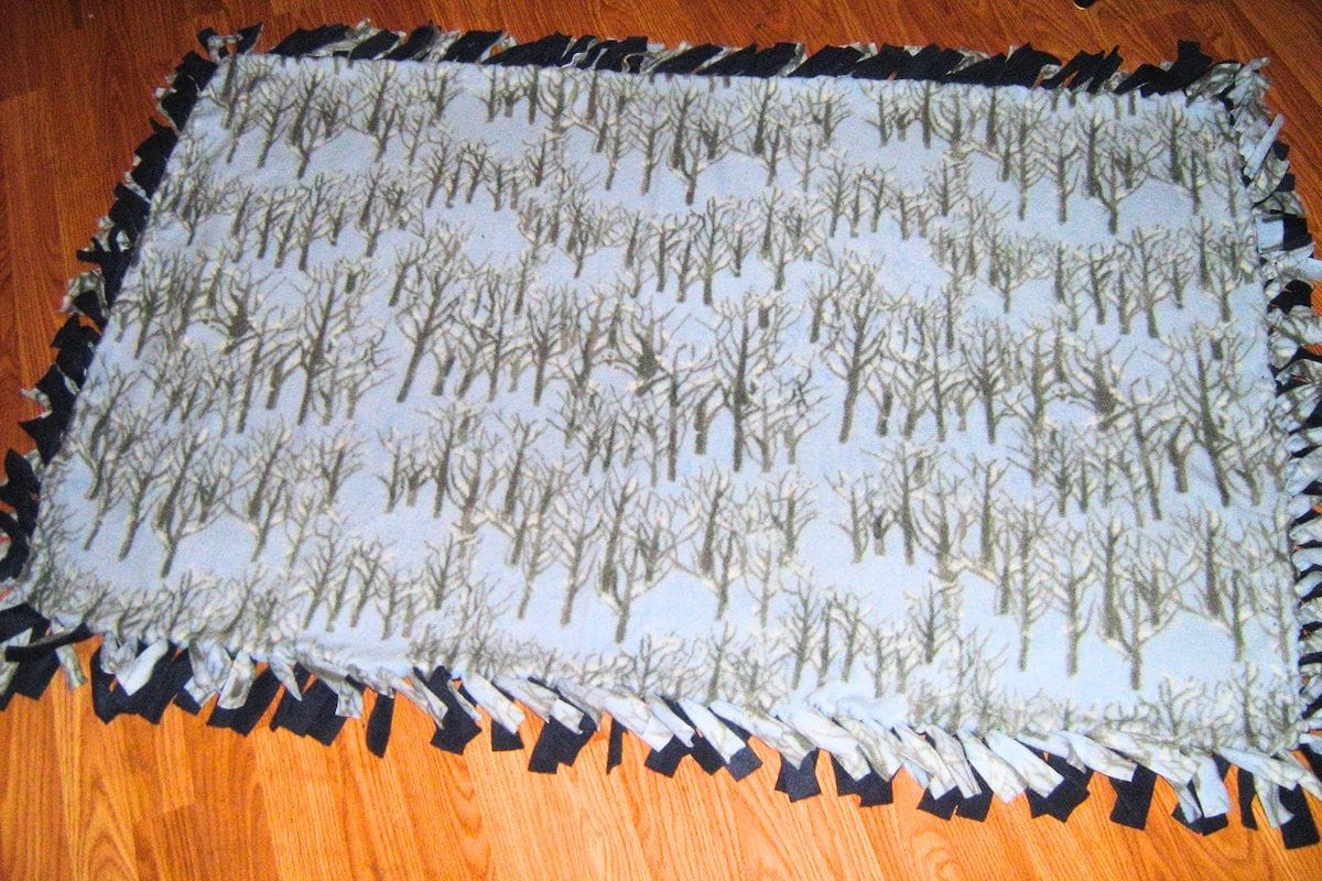Finished no sew fleece blanket with a pale winter forest print featuring rows of bare trees across the fabric. The blanket has contrasting black and patterned fleece fringe tied around the edges.
