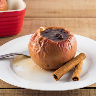 Baked apple with sugar and cinnamon filling on a white plate next to cinnamon sticks.