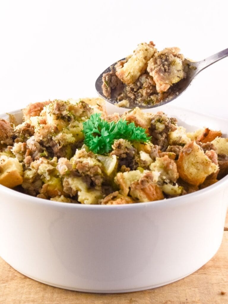 Sausage and herb stuffing recipe served in a white casserole dish with chunks of toasted bread sausage celery and herbs. A spoon lifts a serving from the dish to show the hearty texture.