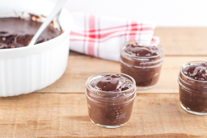 Chocolate pudding served in 3 small mason jars and a large bowl in the background on a wooden table top. A red and white tea towel is laying in the background.