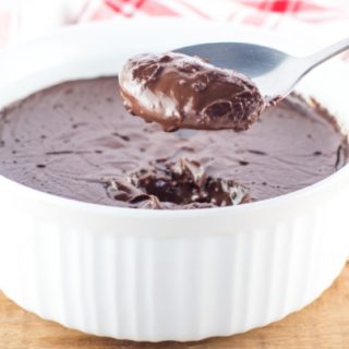 Homemade chocolate pudding in a white bowl on a wooden table. There is a large spoonful of pudding being held over the bowl and a red and white tea towel in the background.