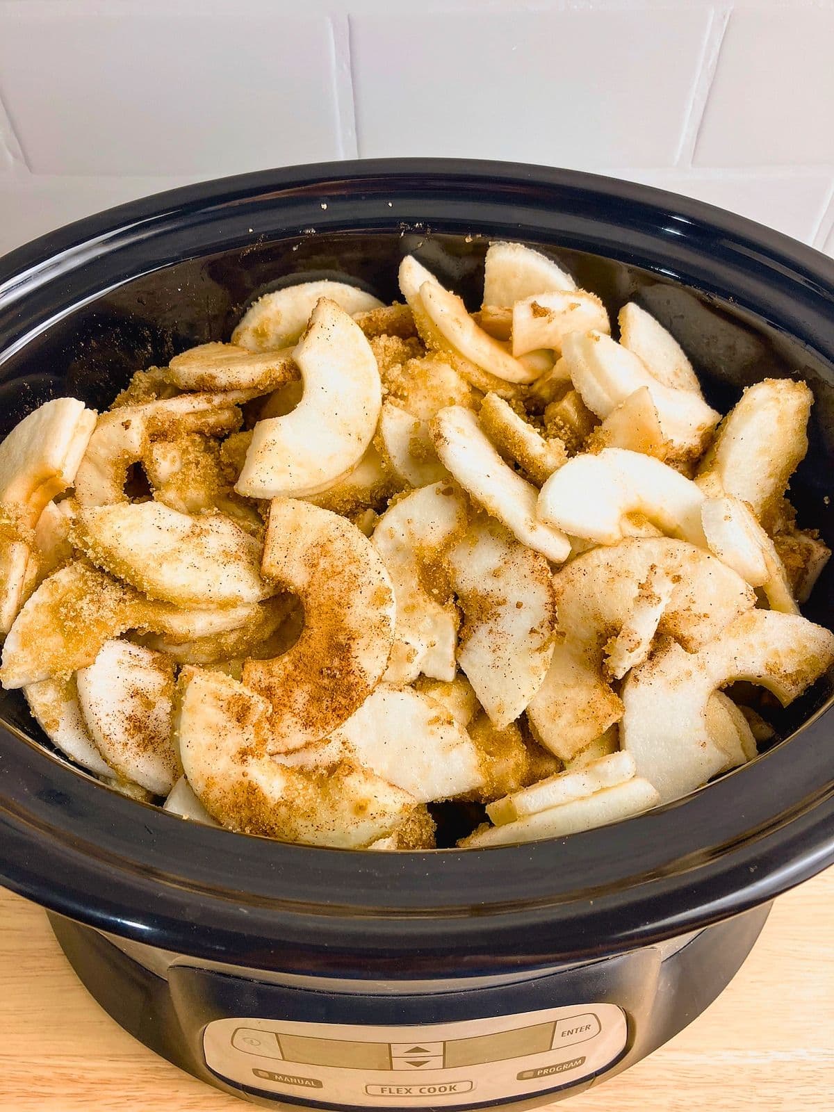 Peeled apple slices coated with brown sugar and warm spices inside a black slow cooker. This step in the slow cooker apple butter recipe shows the apples mixed with the seasonings and ready to begin cooking.