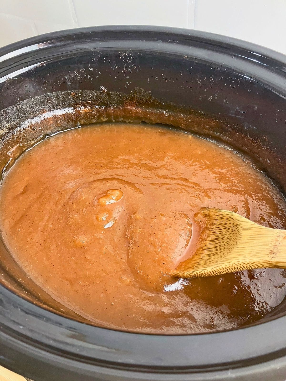 Thick smooth brown apple butter cooking in a black slow cooker with a wooden spoon drawn through the mixture to show its consistency. The slow cooker apple butter recipe has cooked down to a glossy spreadable texture.