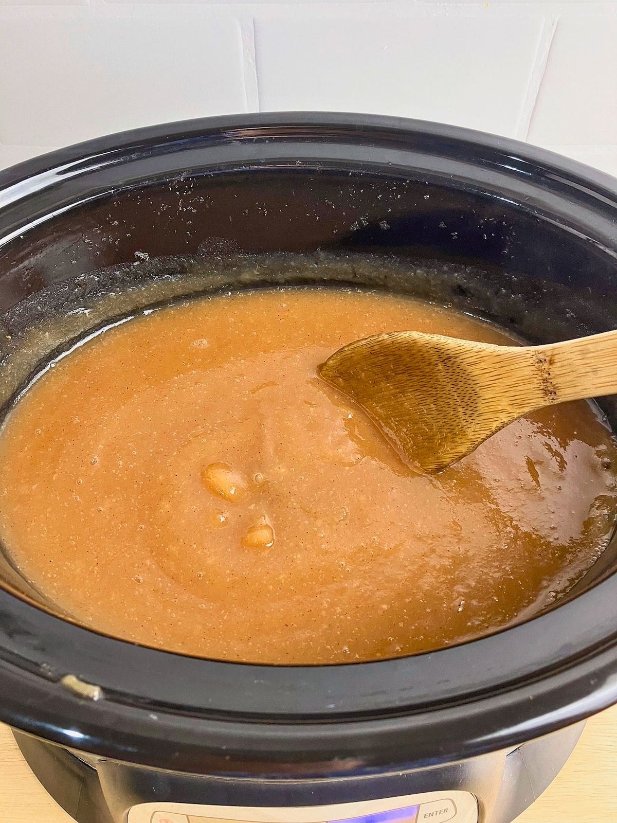 Smooth light brown apple butter cooking in a black slow cooker with a wooden spoon resting in the mixture. This stage of the slow cooker apple butter recipe shows the apples blended smooth and continuing to cook down until thick.