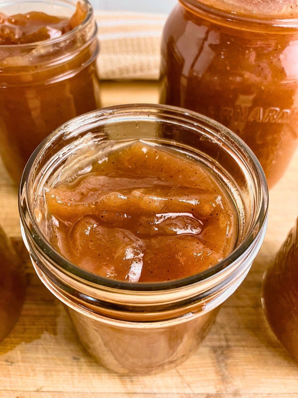 Open glass jars filled with thick glossy brown apple butter arranged on a wooden surface. The finished slow cooker apple butter recipe has a smooth spreadable texture with visible specks of warm spices.