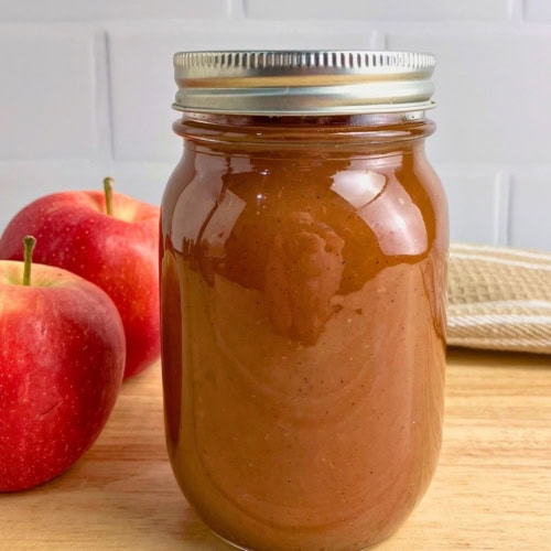 Mason jar filled with smooth brown apple butter on a wooden board beside two red apples. The finished slow cooker apple butter recipe is shown sealed in a jar and ready to store or serve.