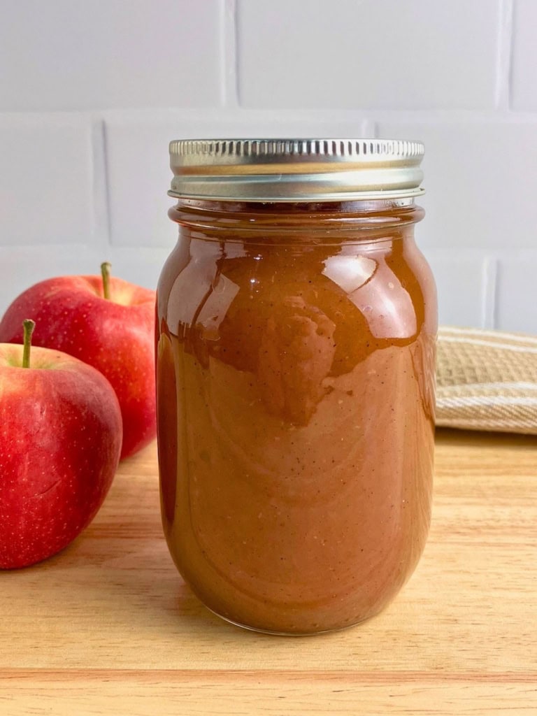 Mason jar filled with smooth brown apple butter on a wooden board beside two red apples. The finished slow cooker apple butter recipe is shown sealed in a jar and ready to store or serve.