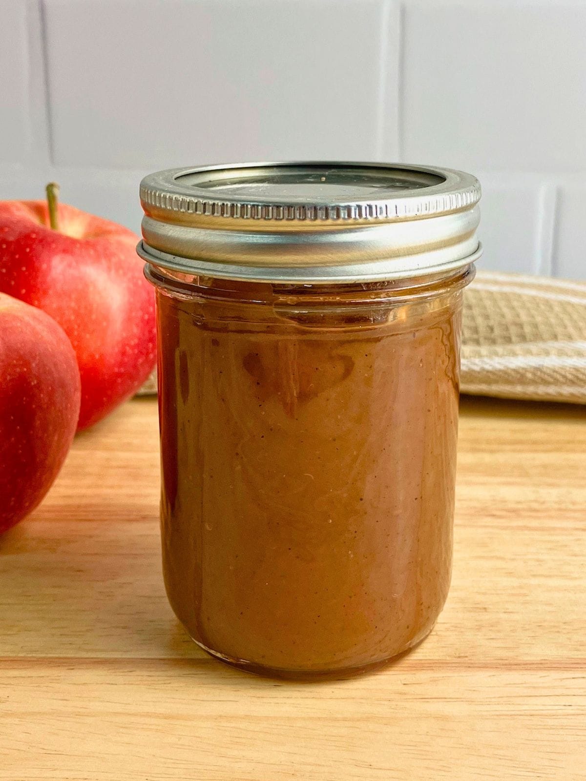 Sealed mason jar filled with smooth brown apple butter on a wooden board with two red apples beside it. The finished slow cooker apple butter recipe is shown ready for storage or serving.