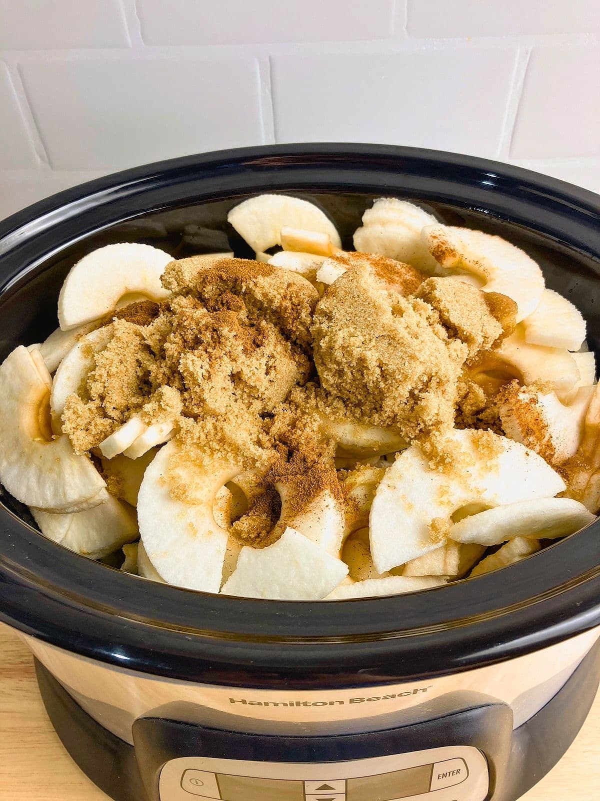 Peeled apple slices in a slow cooker topped with brown sugar and warm spices before cooking.