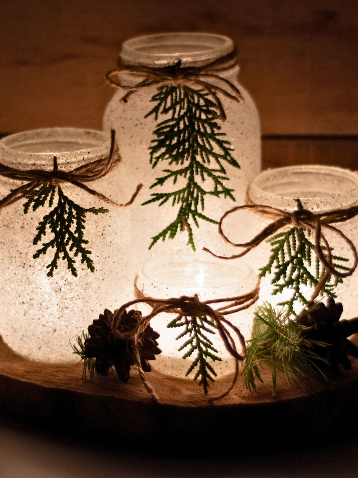 DIY snowy mason jars glow with candlelight and are decorated with evergreen sprigs and rustic twine bows. Pinecones and greenery surround the jars on a wood slice for a cozy Christmas centerpiece.