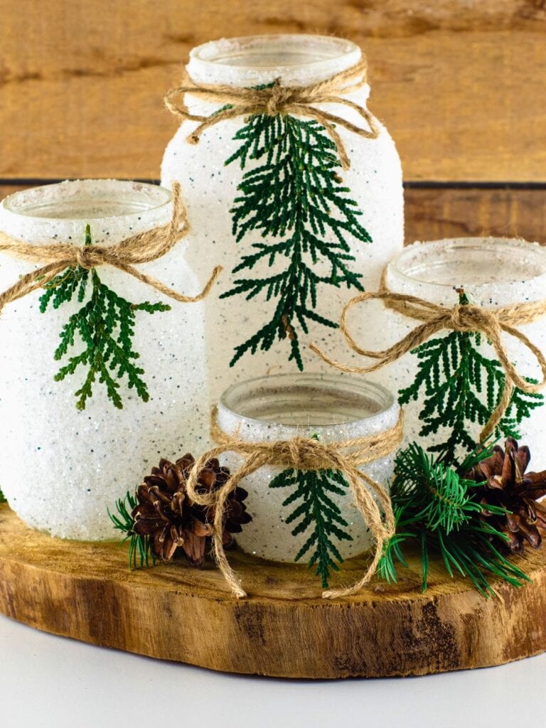 DIY snowy mason jars coated in white glitter are decorated with evergreen sprigs and rustic twine bows. The jars are arranged on a wood slice with pinecones and greenery for a simple Christmas centerpiece.