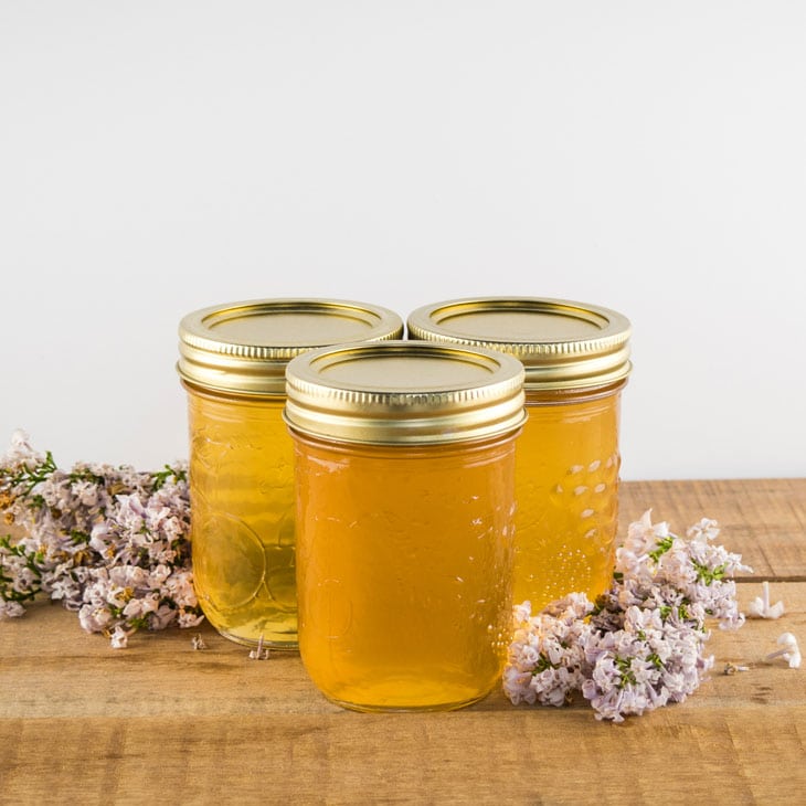 Three jars of homemade lilac flower jelly on a wooden table surrounded by fresh lilac blossoms.