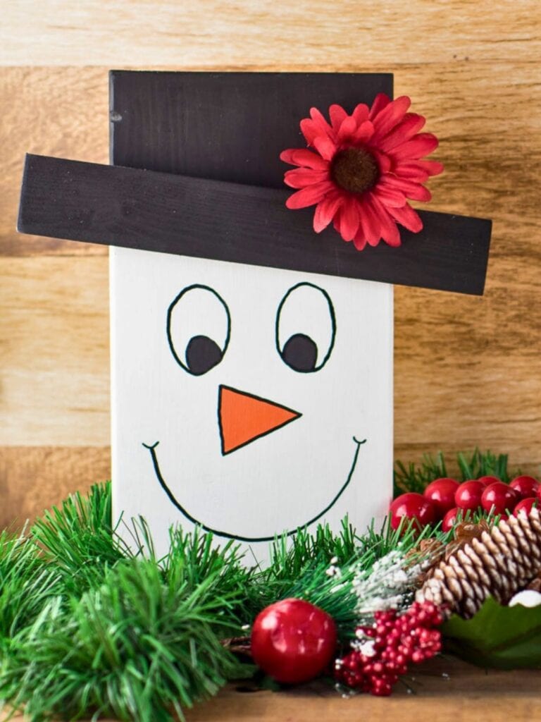 A cute wooden snowman face sitting on a table with a red flower on its hat and surrounded by Christmas greenery and decorations. How to make tabletop wooden snowman faces.