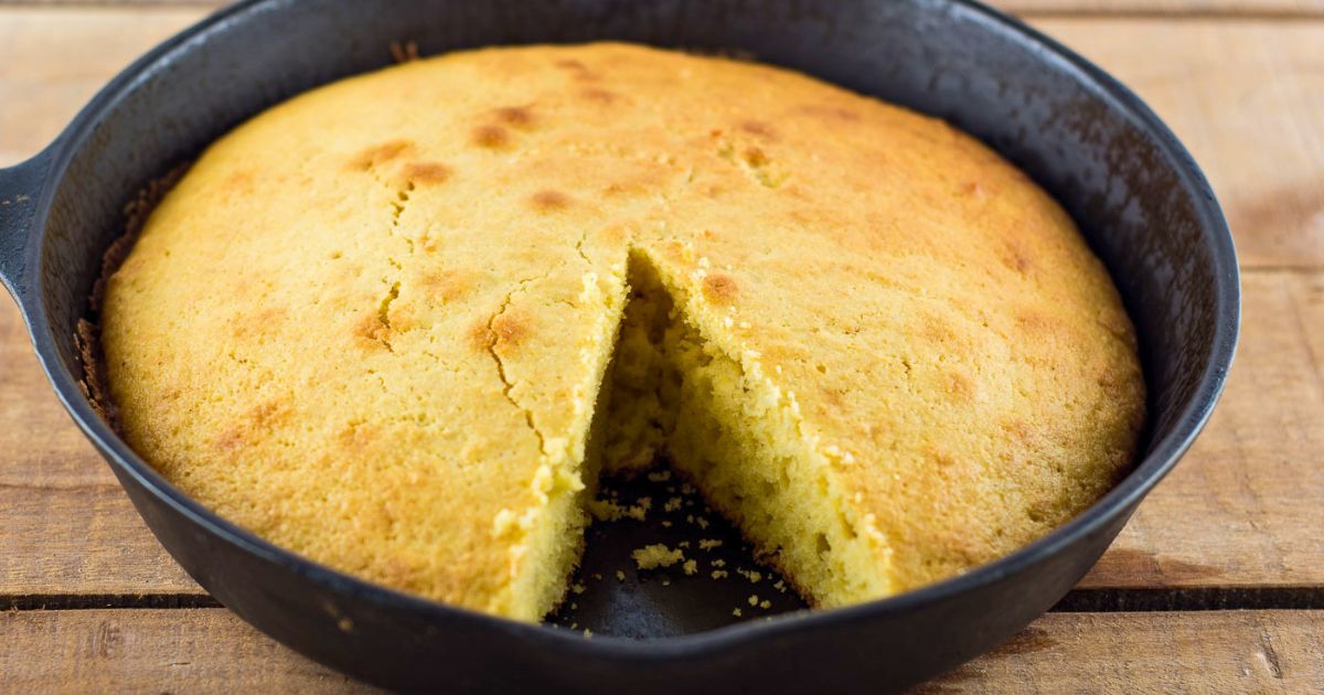 A cast iron skillet of homemade cornbread on a wooden table with a slice removed.