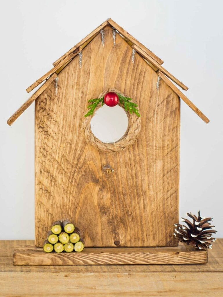 Easy Christmas birdhouse ornament craft made from stained wood with a twine-wrapped circular opening, red berry and greenery accent, mini log stack, and pinecone on a wooden base.