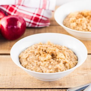Two white bowls filled with homemade apple cinnamon oatmeal on a wooden table next to fresh apples.
