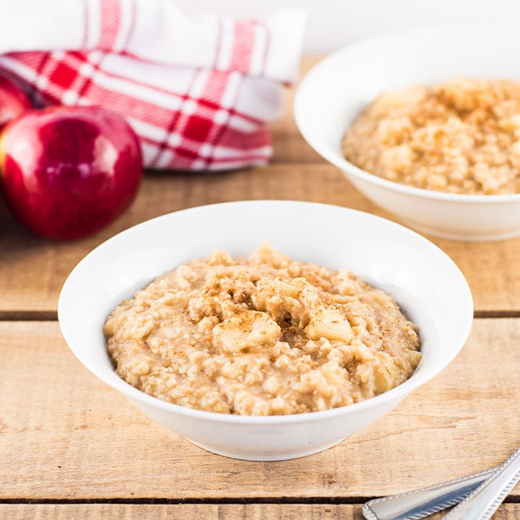 Stovetop Apple Cinnamon Oatmeal Recipe. Warm oatmeal in white bowls sitting on a wooden table with red apples and red and white tea towel in the background.