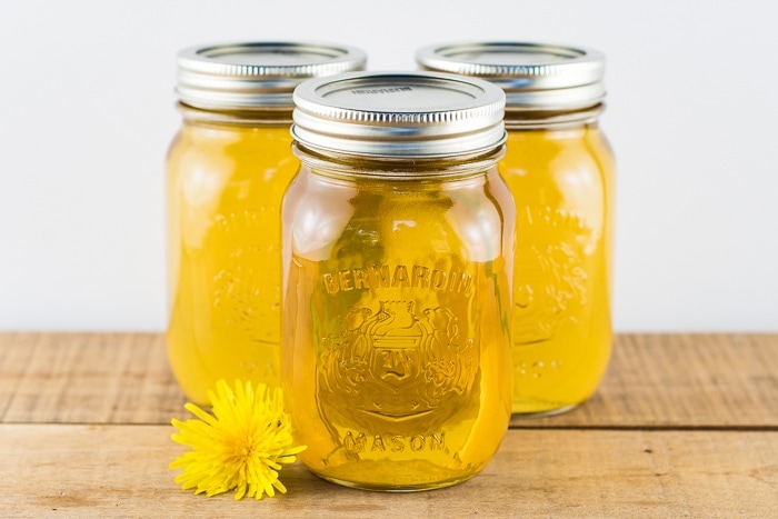 3 pint jars of dandelion syrup canned and sitting on a wooden table with a dandelion flower next to them.