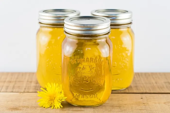 3 pint jars of dandelion syrup canned and sitting on a wooden table with a dandelion flower next to them.