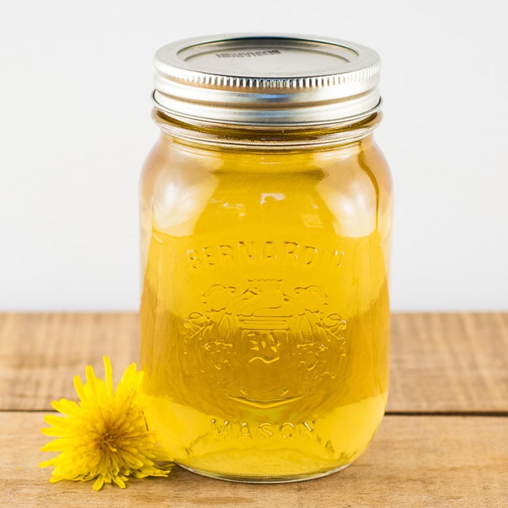 Dandelion syrup in a pint mason jar sitting on a wooden table with a dandelion flower next to it.