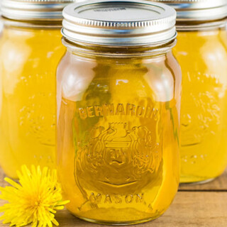 3 jars of dandelion syrup set on a wooden table with a dandelion flower next to them.