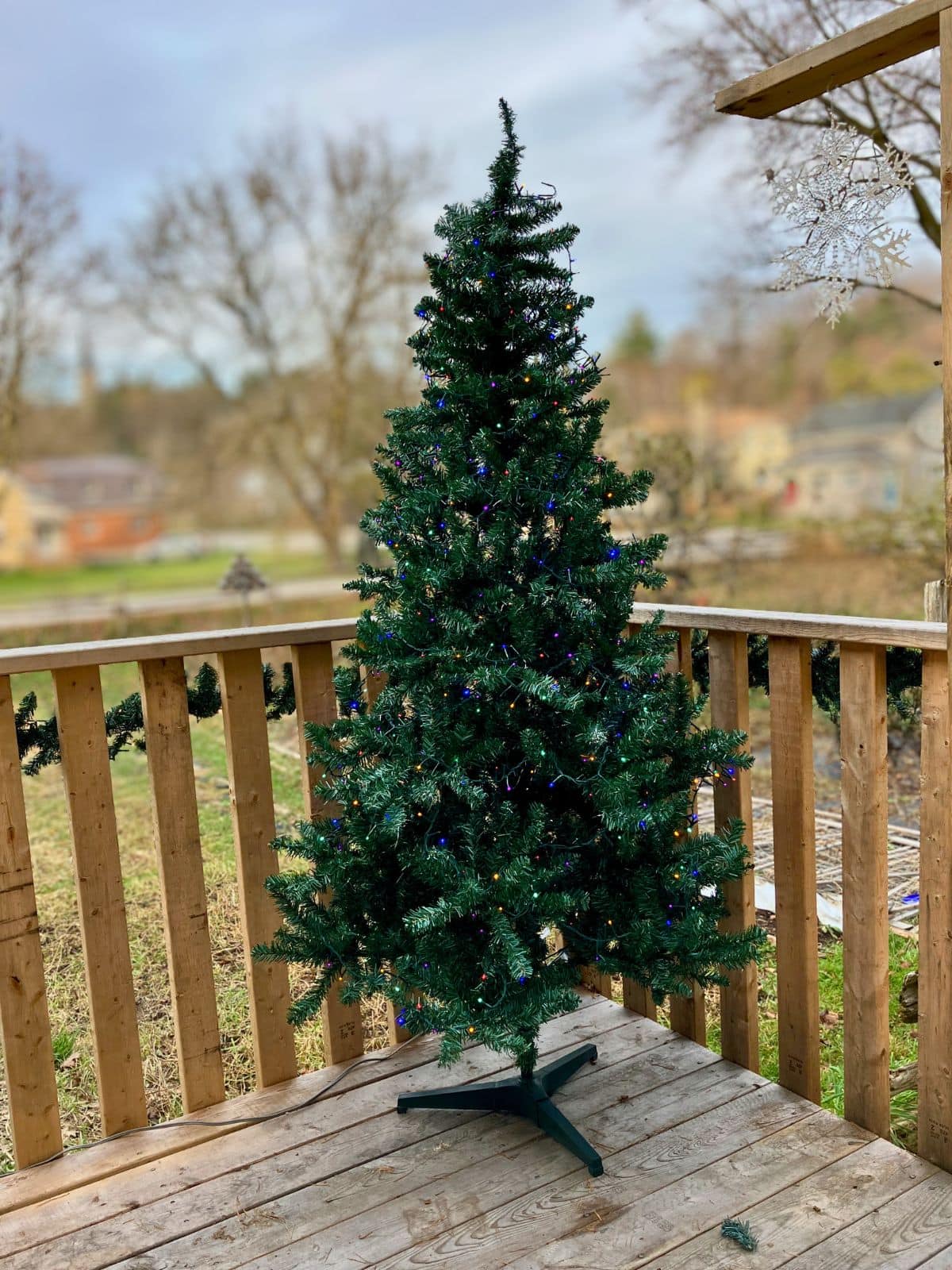 An artificial Christmas tree with multicolored lights stands on a wooden porch outdoors among bare trees and neighboring houses. It shows how a fake Christmas tree can be displayed outside for holiday decorating.