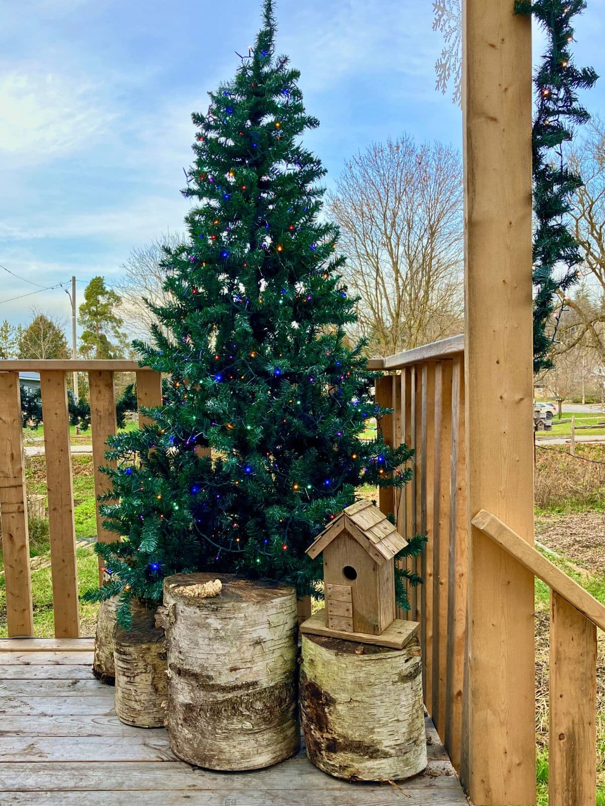 An artificial Christmas tree with multicolored lights stands on a wooden porch beside birch log decorations and a rustic birdhouse. The outdoor setup shows how a fake Christmas tree can be used outside for holiday decorating.