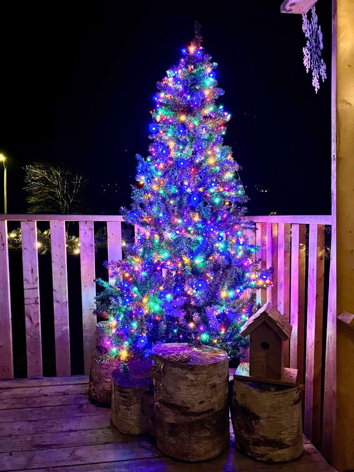 An artificial Christmas tree glows with bright multicolored lights on a wooden porch at night beside birch log decorations and a rustic birdhouse. The outdoor display shows a fake Christmas tree used outside for festive holiday decorating.