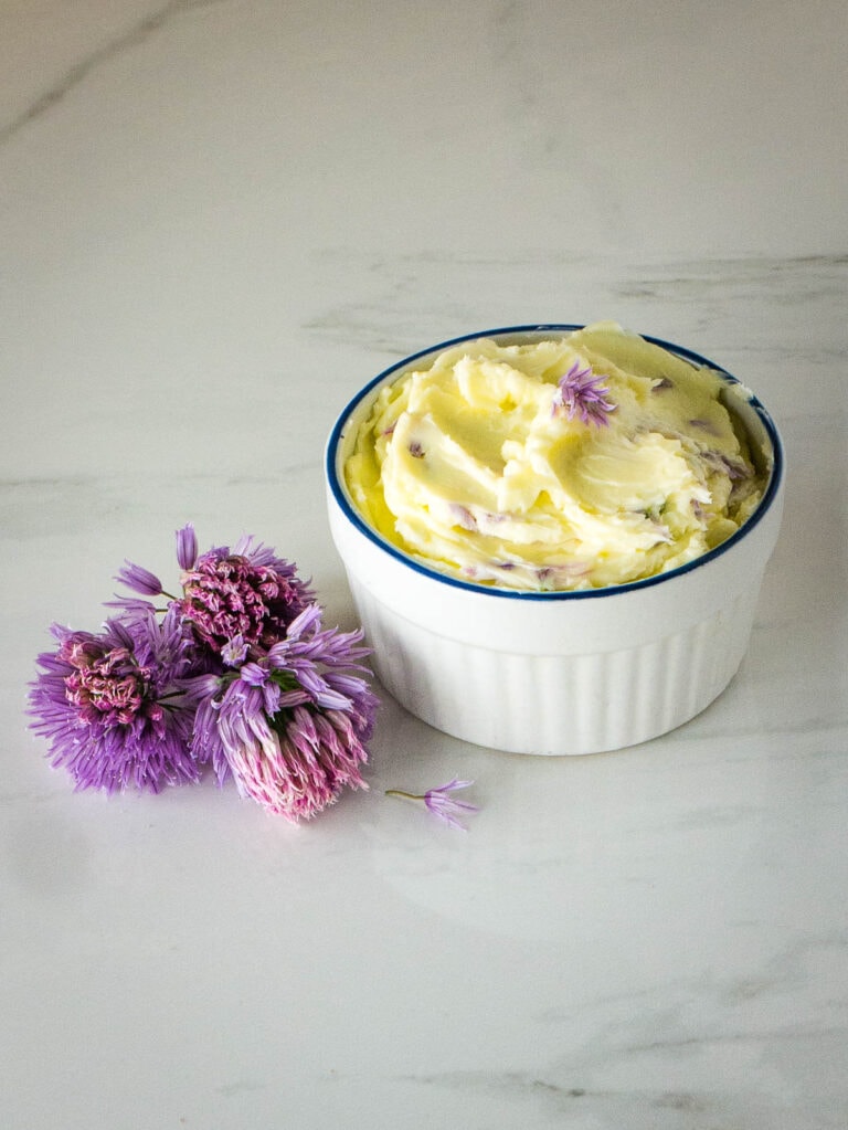 Finished chive blossom butter in a white container on the counter. Next to it are some fresh chive flowers.