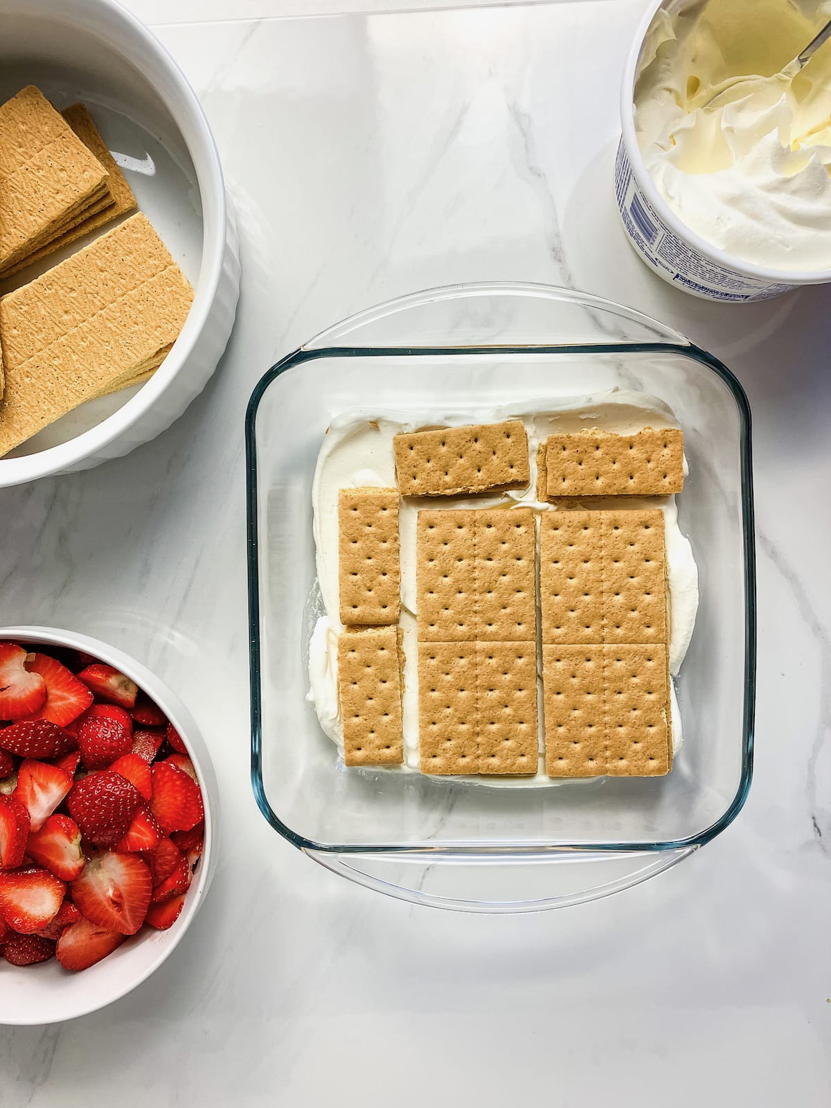 A layer of graham crackers over top of whipped topping in a baking tray. Next to bowls of strawberries, whipped topping, and graham crackers.