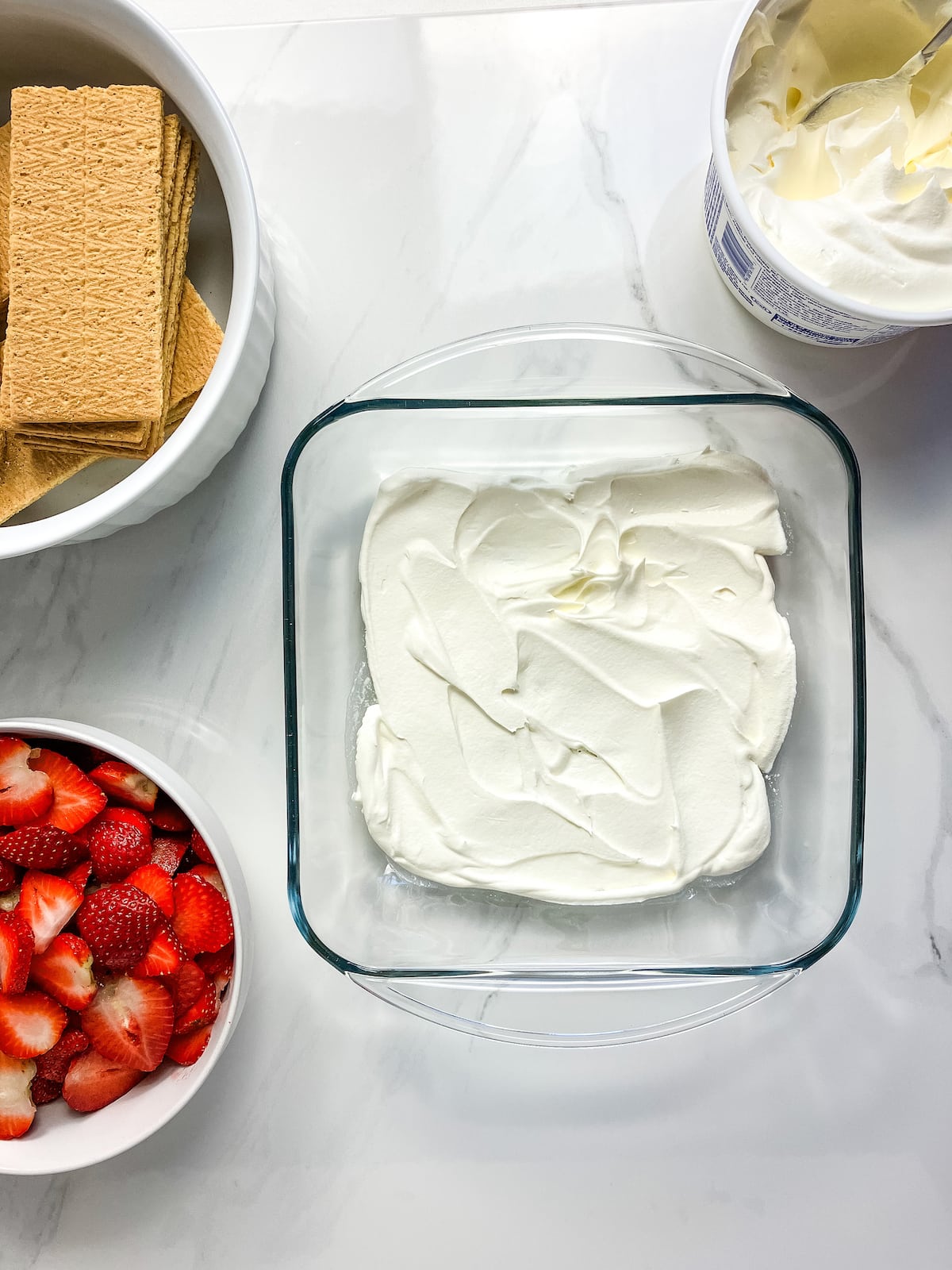 An 8x8 glass baking pan with a layer of whipped topping on the bottom. Next to the pan are bowls of sliced strawberries, graham crackers, and whipped topping.