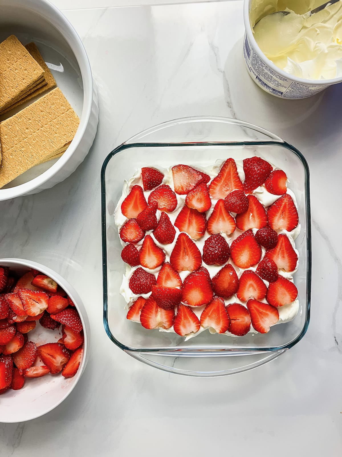 A layer of sliced strawberries on top of the whipped topping in the baking dish. Next to bowls of strawberries, graham crackers and whipped topping.