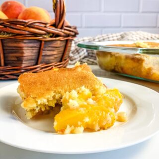 Slice of homemade peach cobbler on a white plate. A baking dish of cobbler and a basket of peaches in the background.