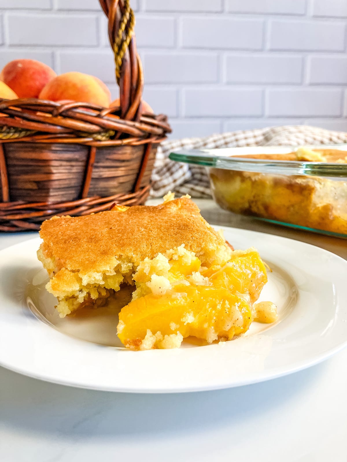 Slice of homemade peach cobbler on a white plate. A baking dish of cobbler and a basket of peaches in the background.