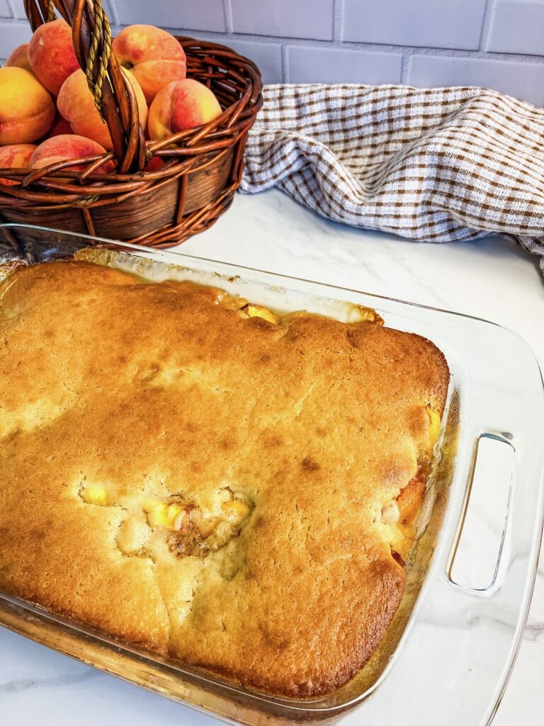 Glass baking dish of homemade peach cobbler. A basket of fresh peaches in the background.