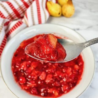 A spoon holding up homemade cranberry pear sauce, over a white bowl of sauce.
