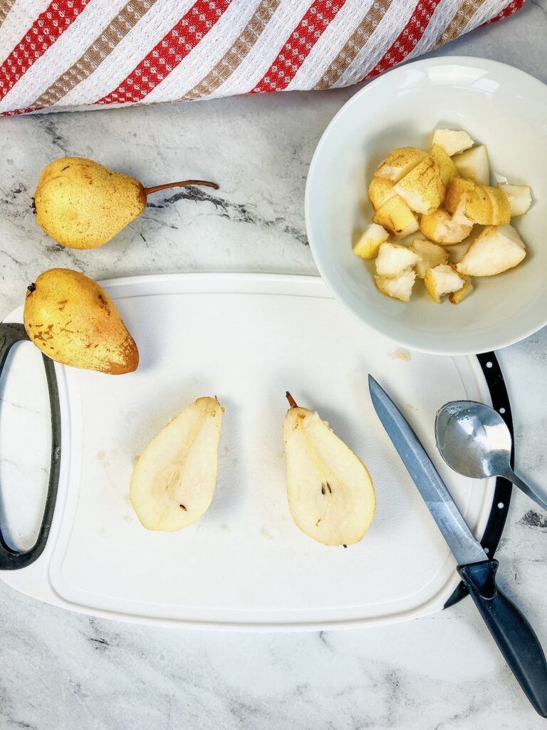 A pear cut in half on a white cutting board. Next to the board is a white bowl of diced pears on the other side 2 whole pears.
