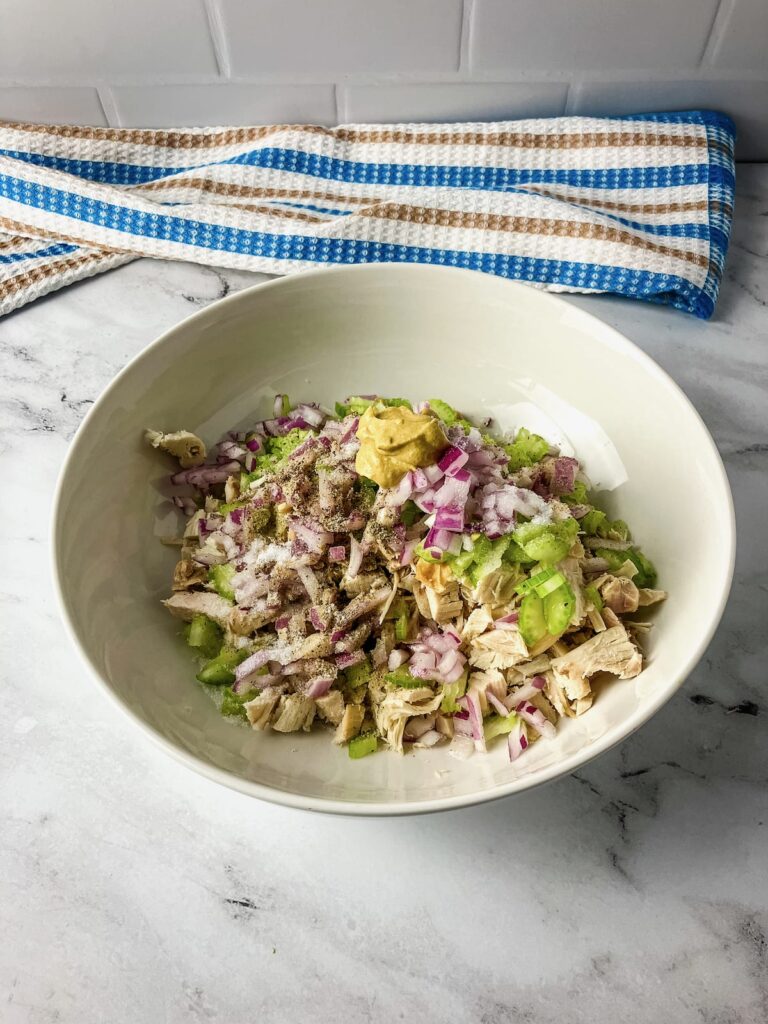 A white mixing bowl with diced turkey, celery, onions, spices and mustard.