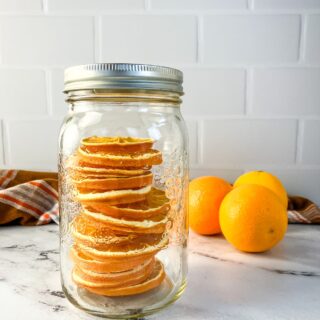 Mason jar filled with dried orange slices on the counter. Fresh oranges in the background.