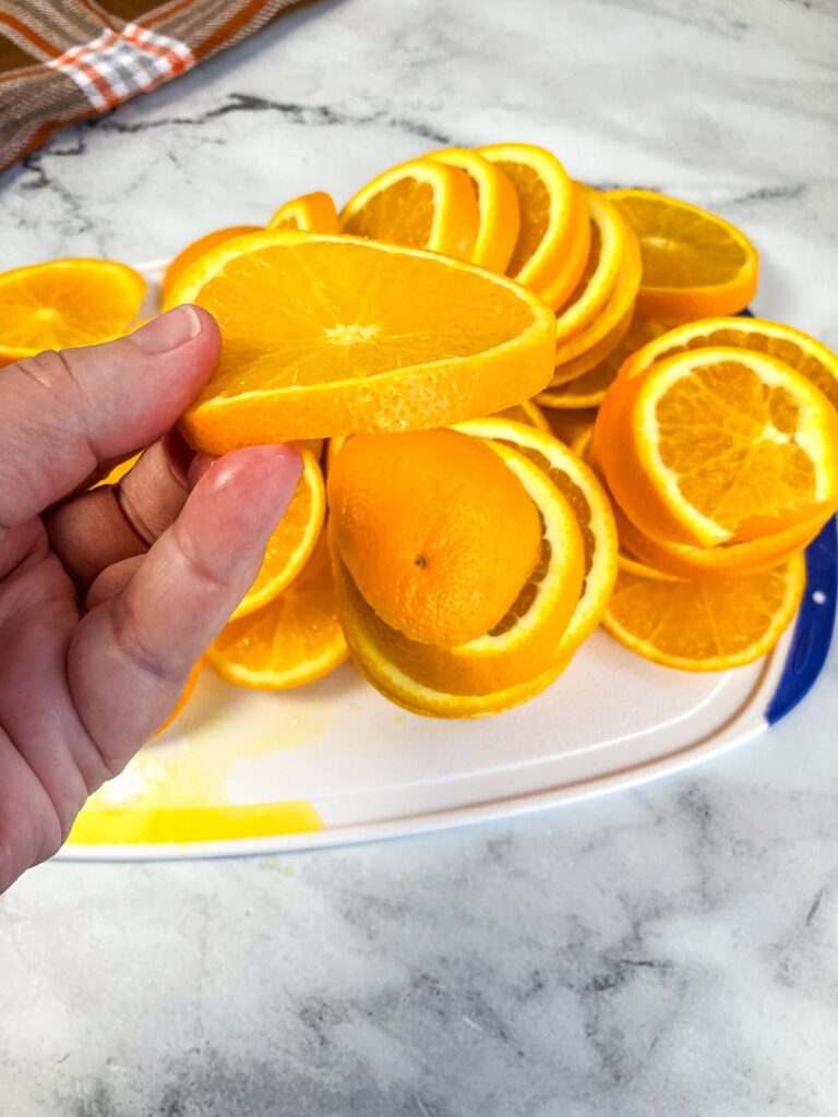 A hand holding a sliced orange in the background is a cutting board filled with sliced oranges.
