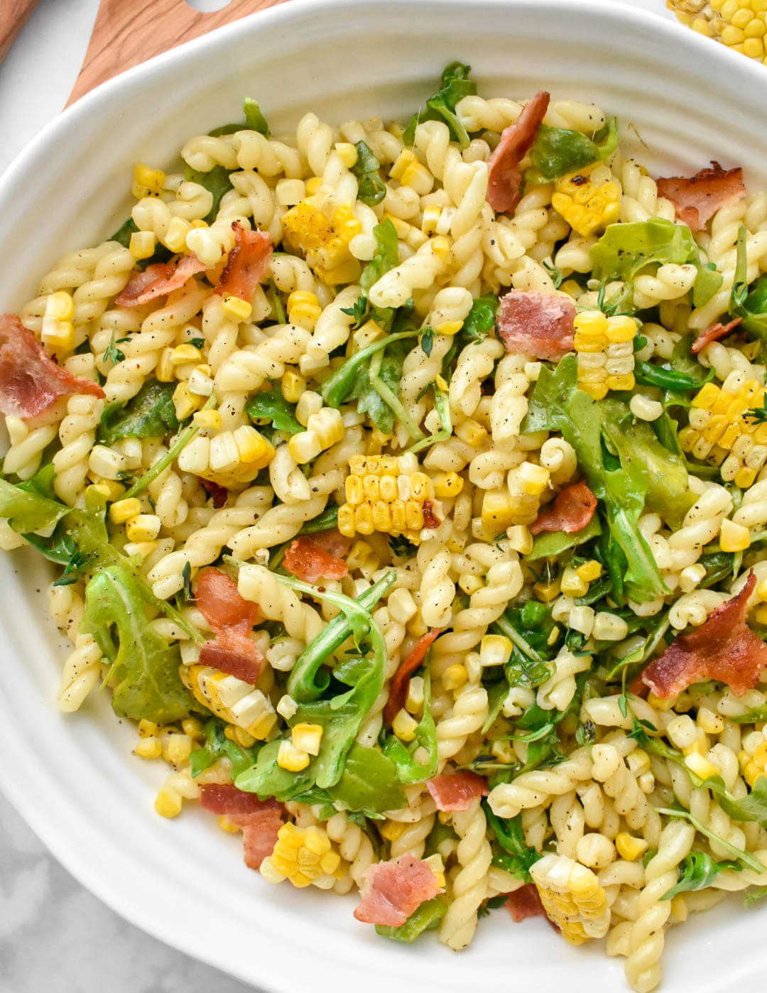Corn, bacon, and arugula pasta salad in a white serving bowl.