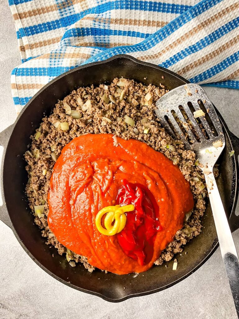Cast iron skillet of ground beef, onion, tomato sauce, ketchup, mustard, salt, and pepper for a sloppy joe filling.