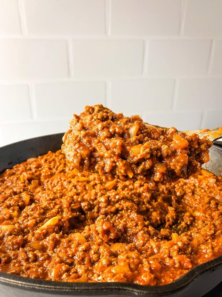 A serving spoon holding up homemade sloppy joes filling mixture in a cast iron skillet.
