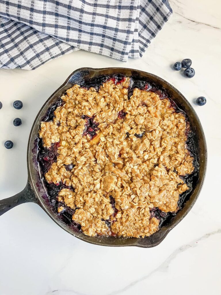 Warm blueberry peach crisp in a skillet with fresh blueberries on the counter around it.