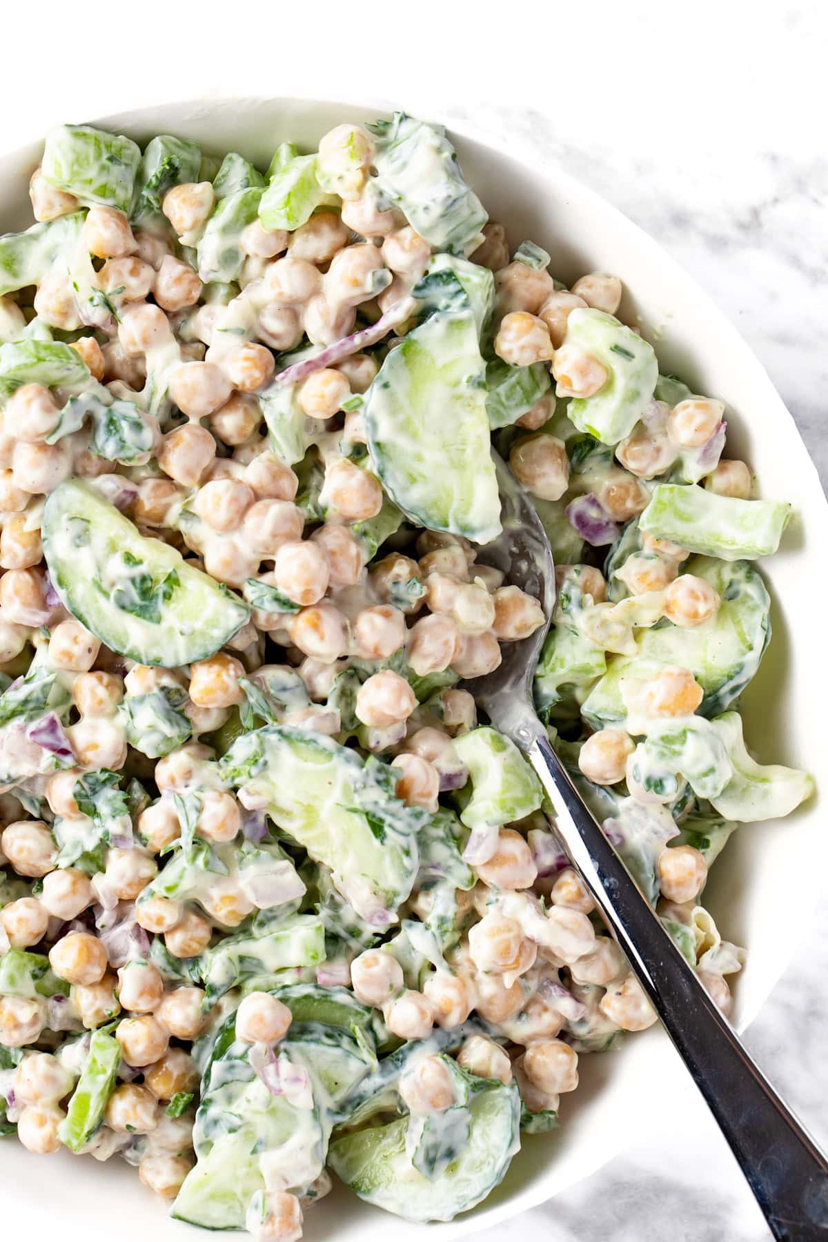 A white bowl filled with chickpeas, cucumbers, and veggies tossed in a creamy dressing.