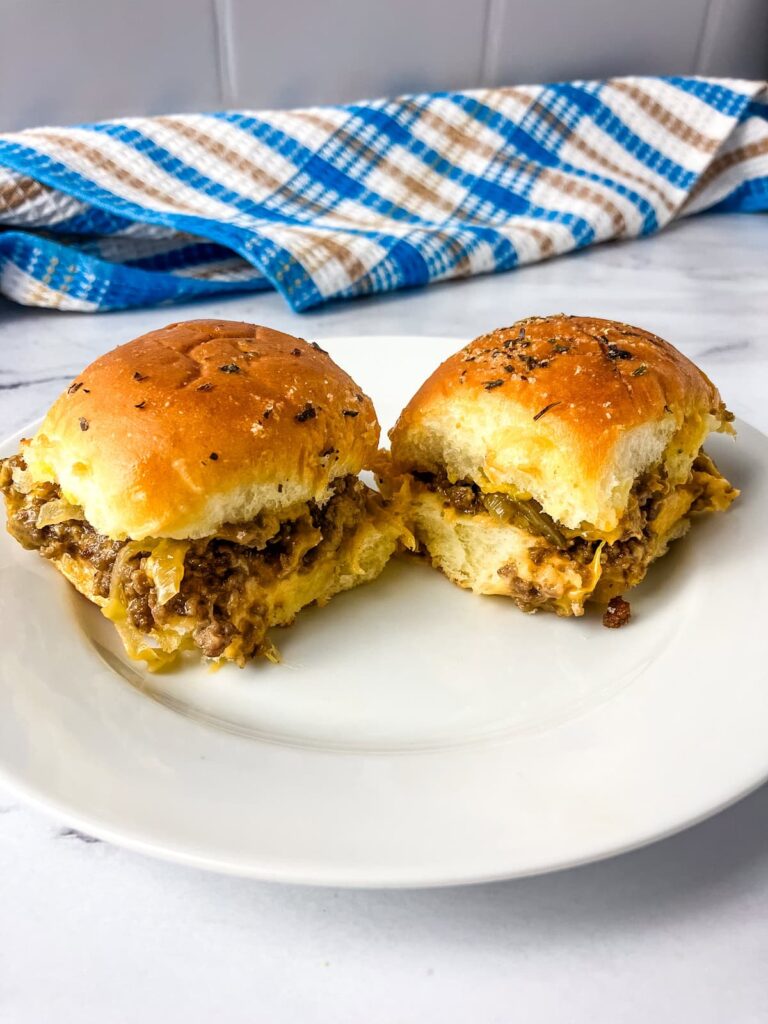 Two homemade cheeseburger sliders on a white plate sitting on a counter with a blue and brown tea towel in the background.