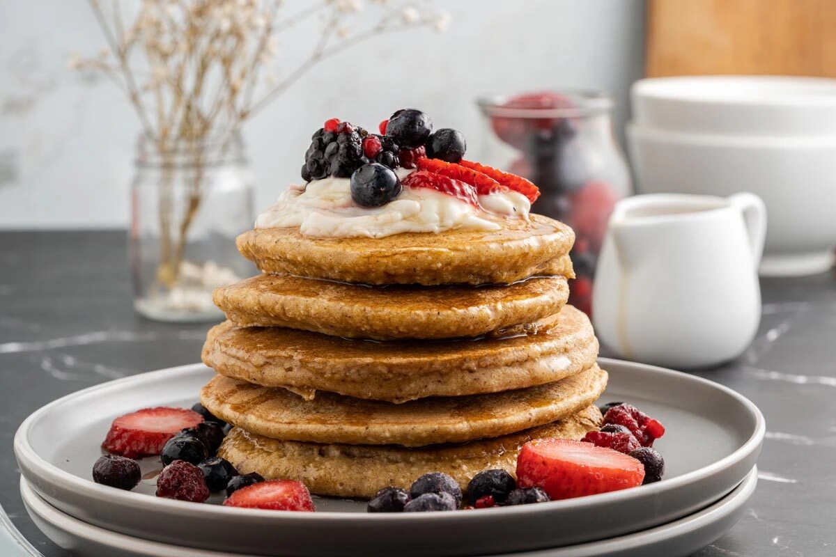 Oat flower pancakes stacked on a plate and topped with fresh fruit.