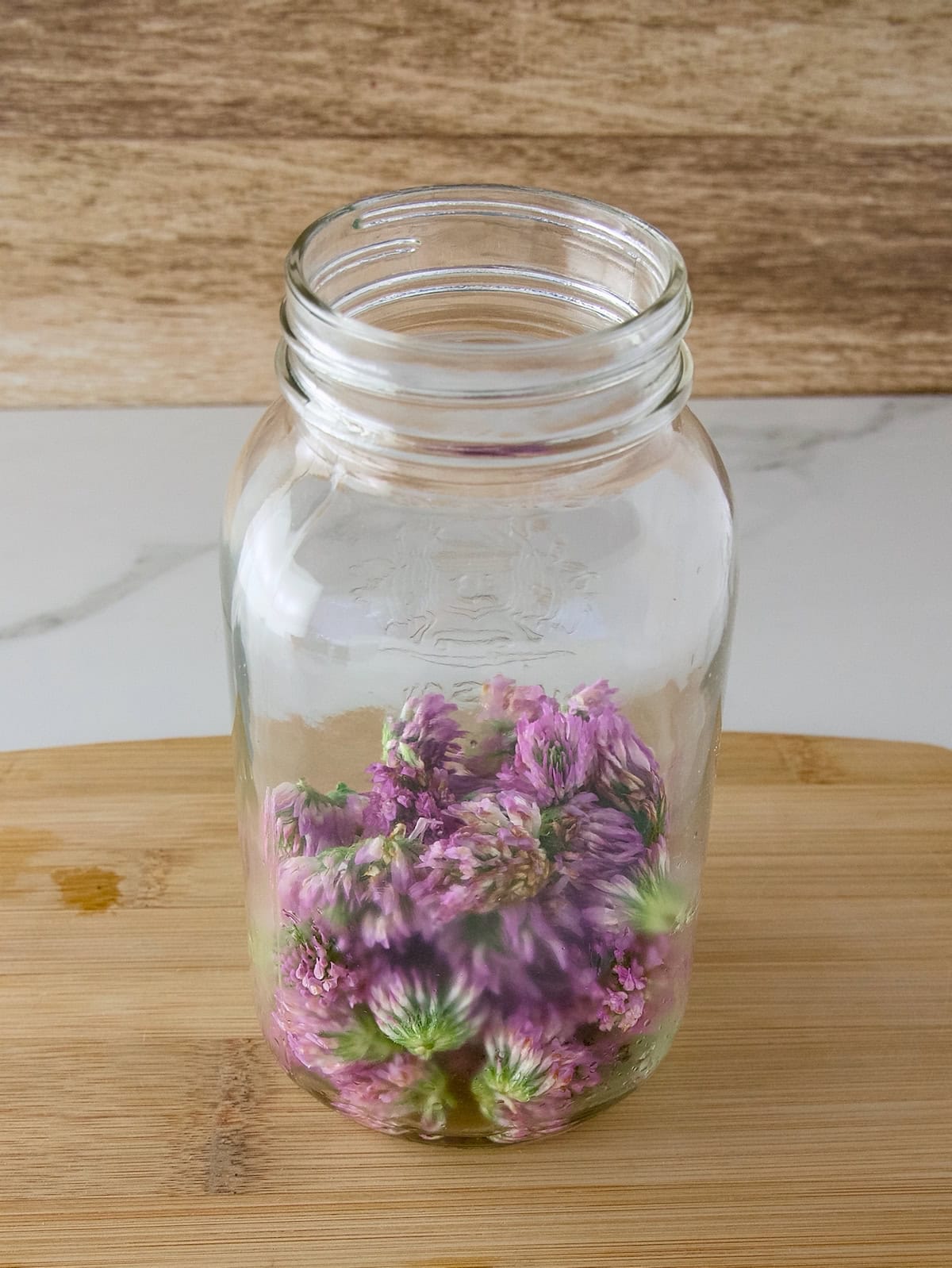 A quart-sized mason jar half filled with red clover flowers, sitting on a wooden cutting board.