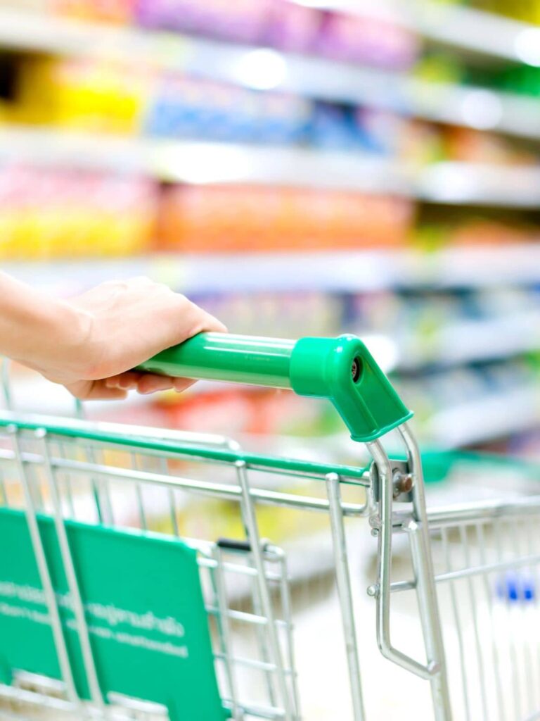 The green handle of a shopping cart is being held as colorful products line the shelves. She is preparing to save money on grocery shopping.